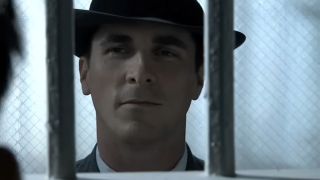 Christian Bale with a fedora on looking through the bars of a jail cell in Public Enemies