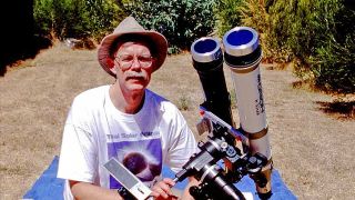 a man in a wide-brimmed hat poses next to a telescope outside on a sunny day