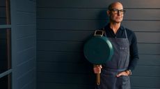 Stanley Tucci in blue apron holding a teal pan against a blue wood panelled backdrop