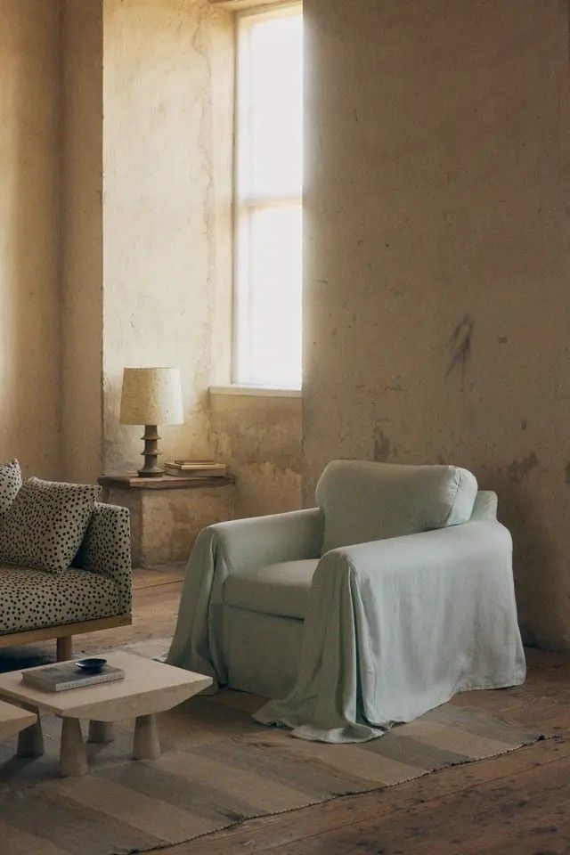 Image of a vintage living room with unfinished-looking plaster walls, a light blue accent chair with a draping cover on it, a blue and white striped rug, and a polka dot sofa in view.