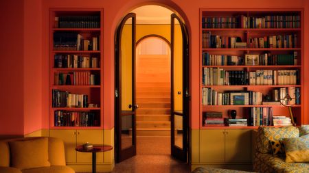 Library with warm red and mustard walls, built-in shelving, arched glazed doors to hallway and patterned yellow and blue sofa