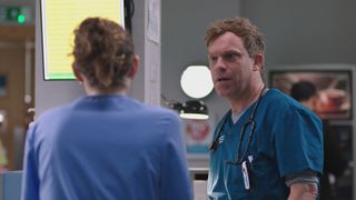 Dylan Keogh in blue scrubs talks to Jodie Whyte in lighter blue scrubs during a quiet moment in Holby ED.