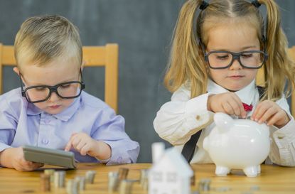 Two toddlers are dressed as adults wearing business attire featuring suspenders and a bow tie. Both of them wear thick rimmed eye glasses and are playing with coins, a piggy bank, money, and 