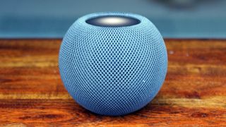 En blå Apple HomePod mini står på ett träfärgat bord mot en suddig, blå bakgrund.