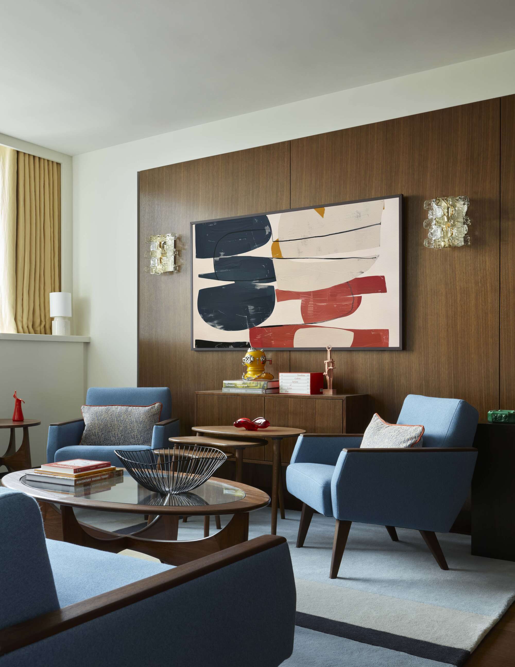 mid-century modern style living room with timber panelled wall, console, nesting tables, two blue armchairs, a blue sofa, a glass and timber round coffee table styled with basket and books