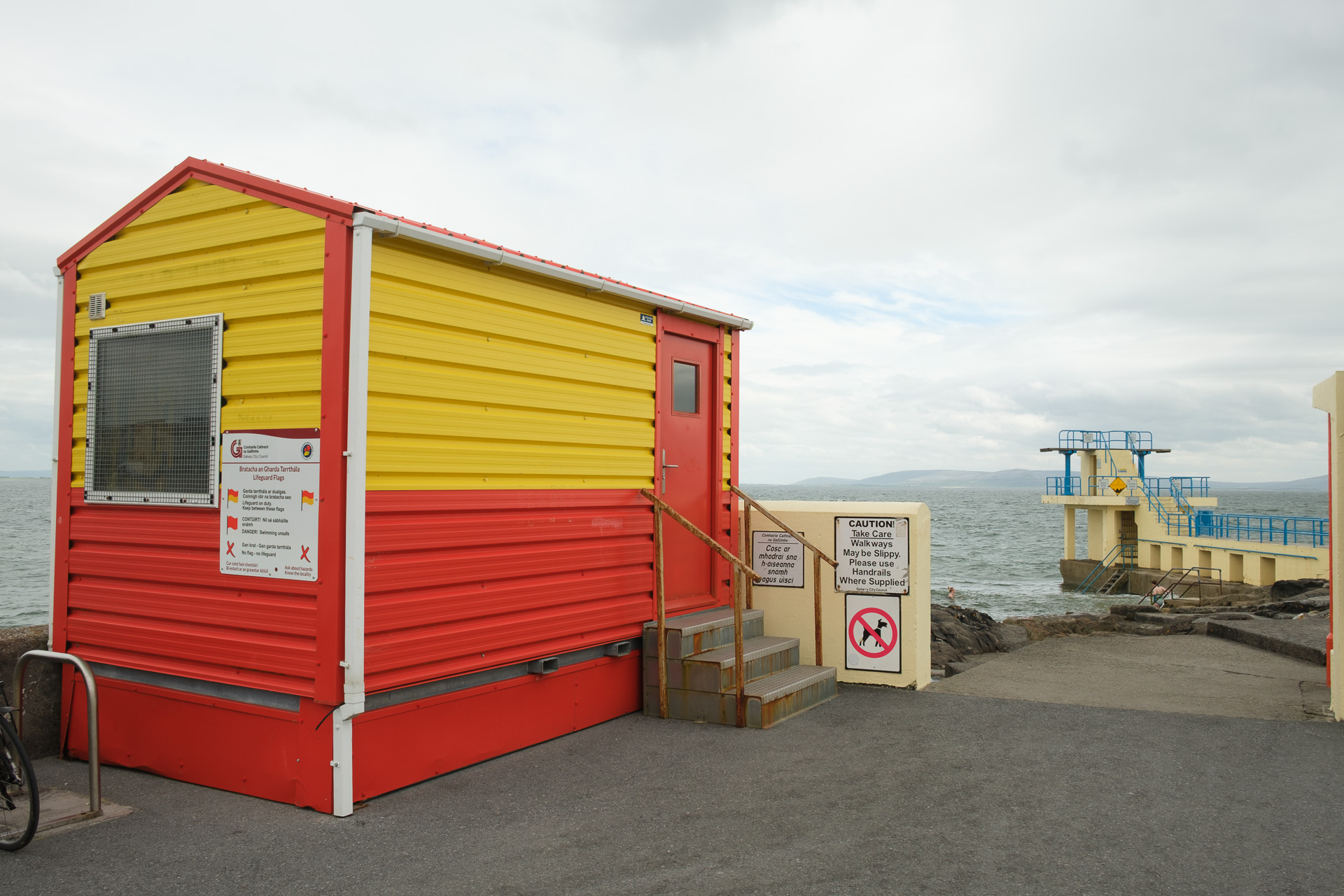 A red and yellow lifeguard hut stands beside the sea, with a cream diving tower visible in the background.