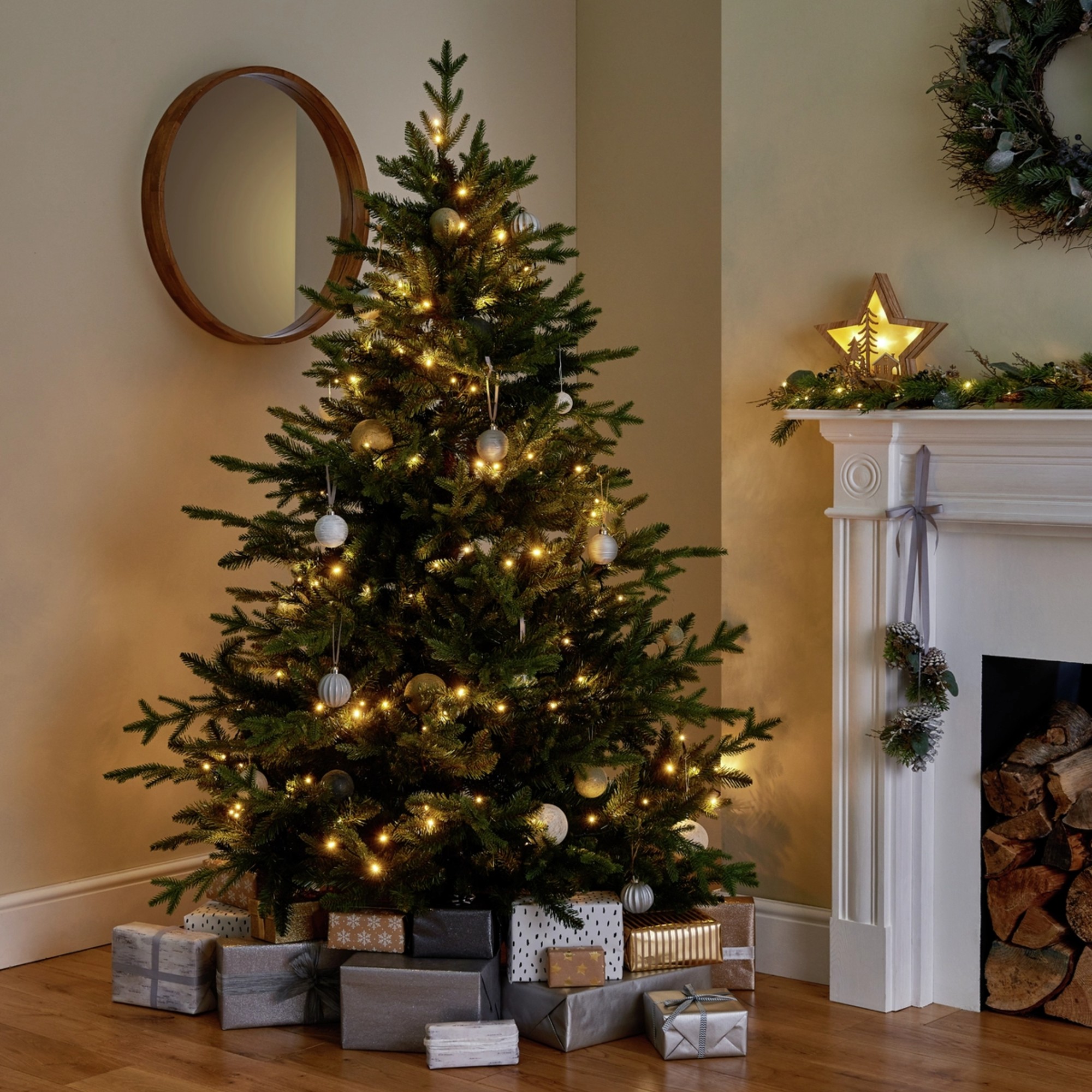 Habitat 6ft Mixed Tip Upswept Christmas Tree next to a white fireplace with light and presents underneath