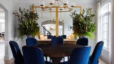 Modern dining room with royal blue upholstered chairs, double wall mirrors, large foliage displays, round dining table, and brass circular feature pendant overhead.
