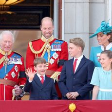 King Charles and Prince William wear red military uniform, Prince Louis and Prince George wear navy suits with red ties, while Princess Kate and Prince Charlotte wear turquoise dresses on the Buckingham Palace balcony