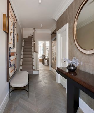 neutral hallway with textured wallpaper, statement artwork, staircase with neutral runner, dark wood console and round mirror
