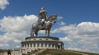 A large silver statue of Genghis Khan sitting on a horse and drawing his sword. In the background are a blue sky and clouds.