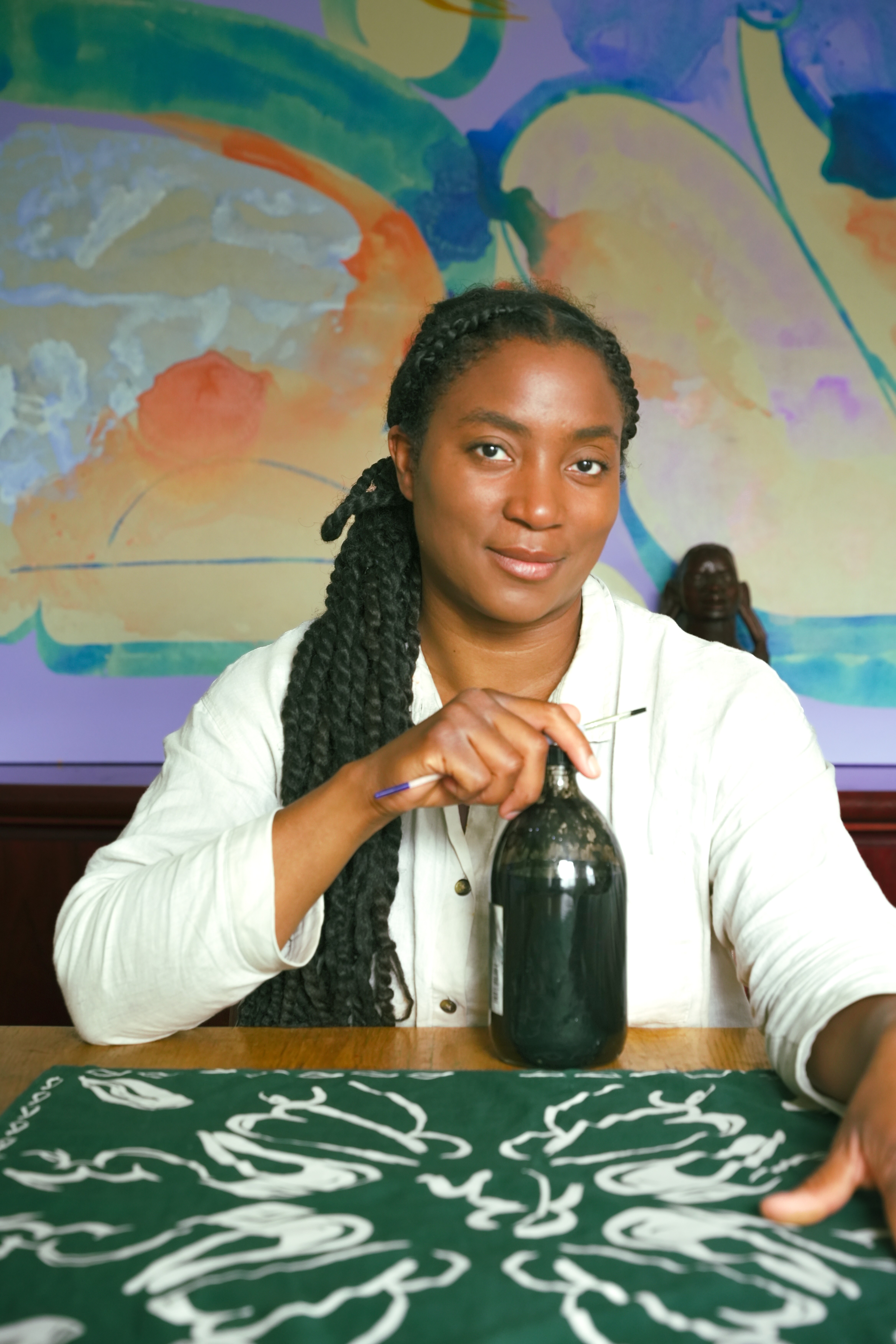A young woman with braided hairs gathered in a long black pony tail resting on her right shoulder sits at a wooden desk while holding a black glass bottle in her right hand and a black-and-white print resting on the table in front of a colorful painting.