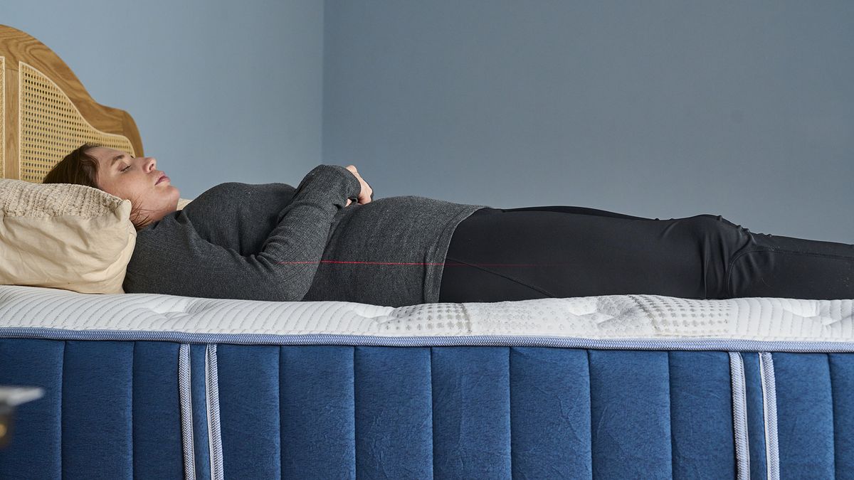 Our mattress tester wearing a dark grey long-sleeved shirt and black leggings is lying on their back on the white and navy Bear Elite Hybrid mattress. A thin red laser line is projected across their midsection to show the straight alignment of the spine and the mattress's support level.