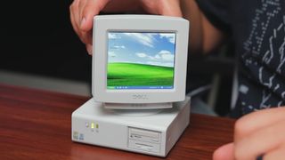 A tiny PC sitting on a wooden desk with its screen tilted to one side, displaying what looks like Windows XP