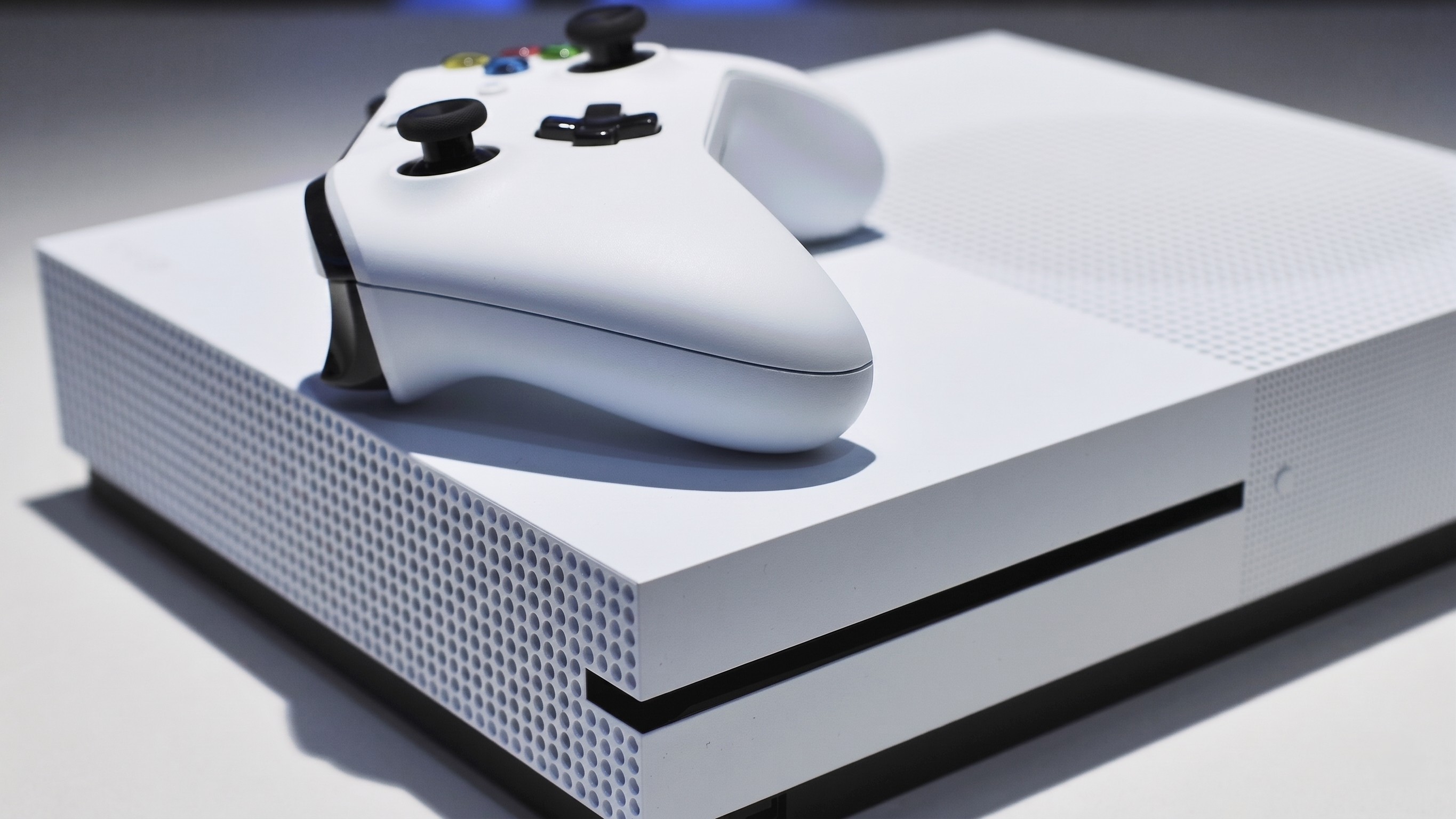 A white Xbox One S showing its disc drive and a matching white Xbox One controller on top.