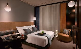 Interior view of a room at Nobu Shoreditch, London, UK featuring brown flooring, grey, blue and black walls, a wooden panel, sphere pendant lights, a bed with grey and white pillows and linen along with bolster pillows, a throw and a tray with items on top, a brown chair with a patterned cushion and a round black table with food and drink on top