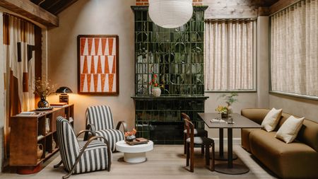 Living area of Mason Fifth Belsize Park with green tiled fireplace, red and pink geometric art, two striped armchairs, brown sofa with dining table and chairs, white coffee table and wooden unit