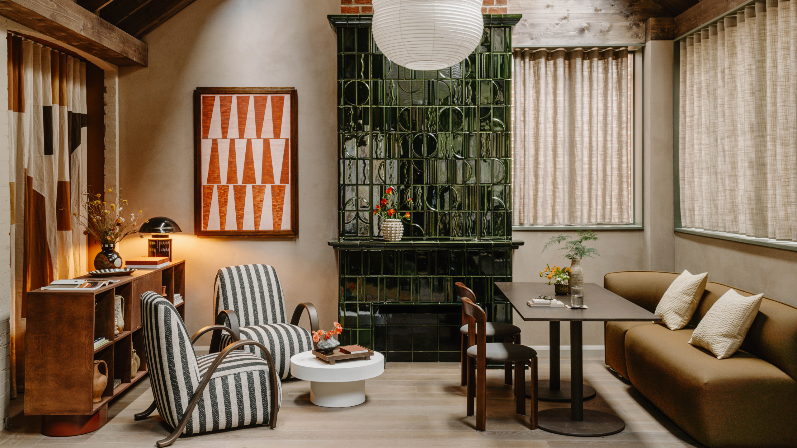 Living area of Mason Fifth Belsize Park with green tiled fireplace, red and pink geometric art, two striped armchairs, brown sofa with dining table and chairs, white coffee table and wooden unit