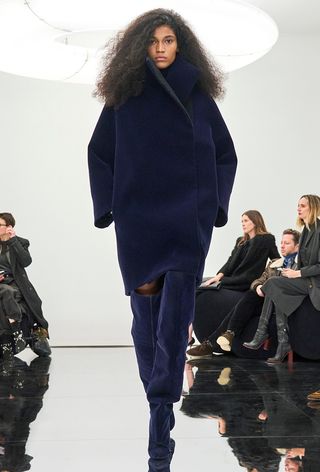 a photo showing some of the best thigh-high boots with a model walking the Alaïa runway show wearing a navy funnel neck knee-length coat styled as a dress paired with matching thigh-high boots