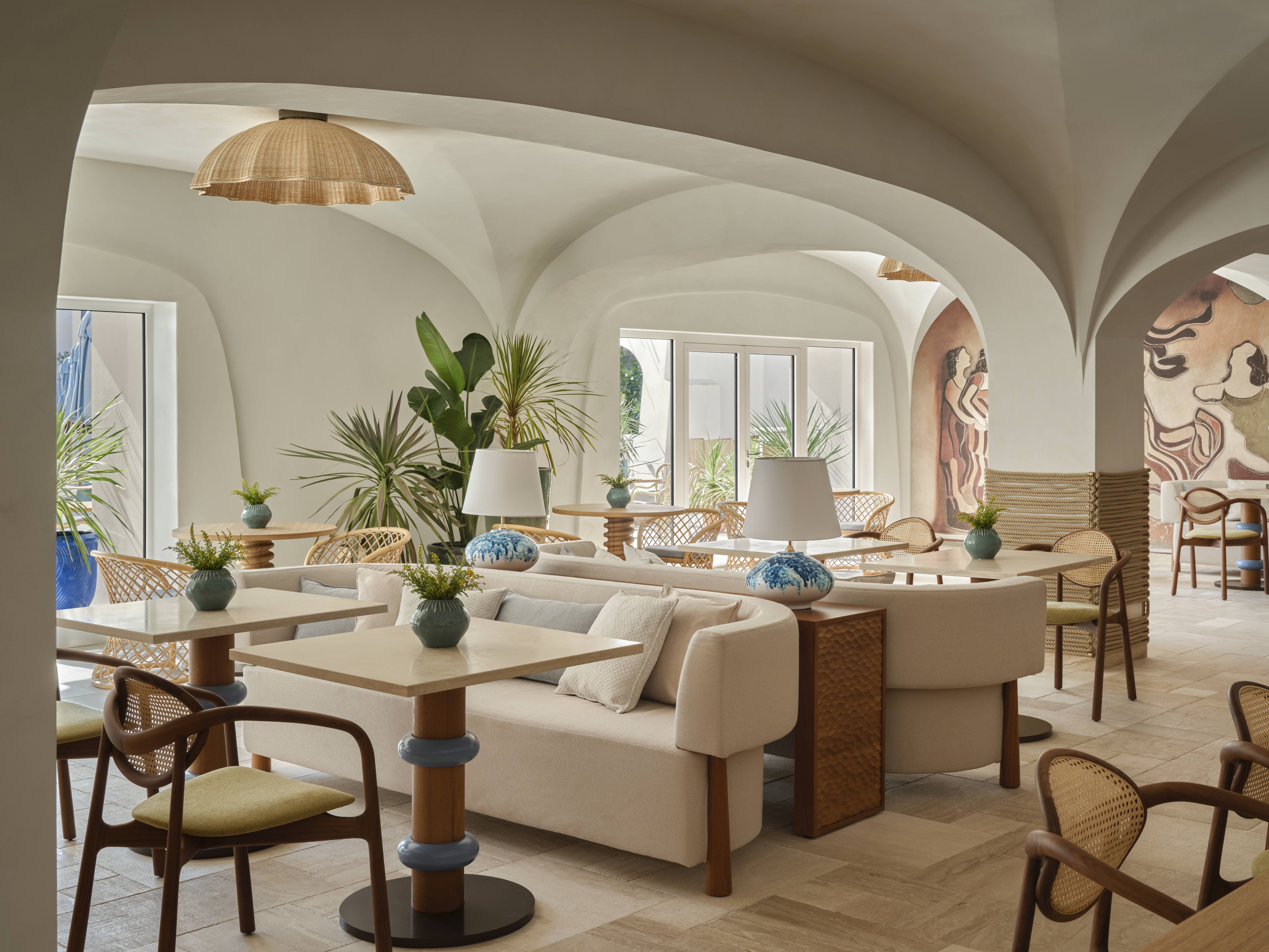A seaside resort features spectacular, arched ceilings painted in a creamy tone, rattan and wooden furniture in playful silhouettes, turquoise accents, and a panoramic pool.