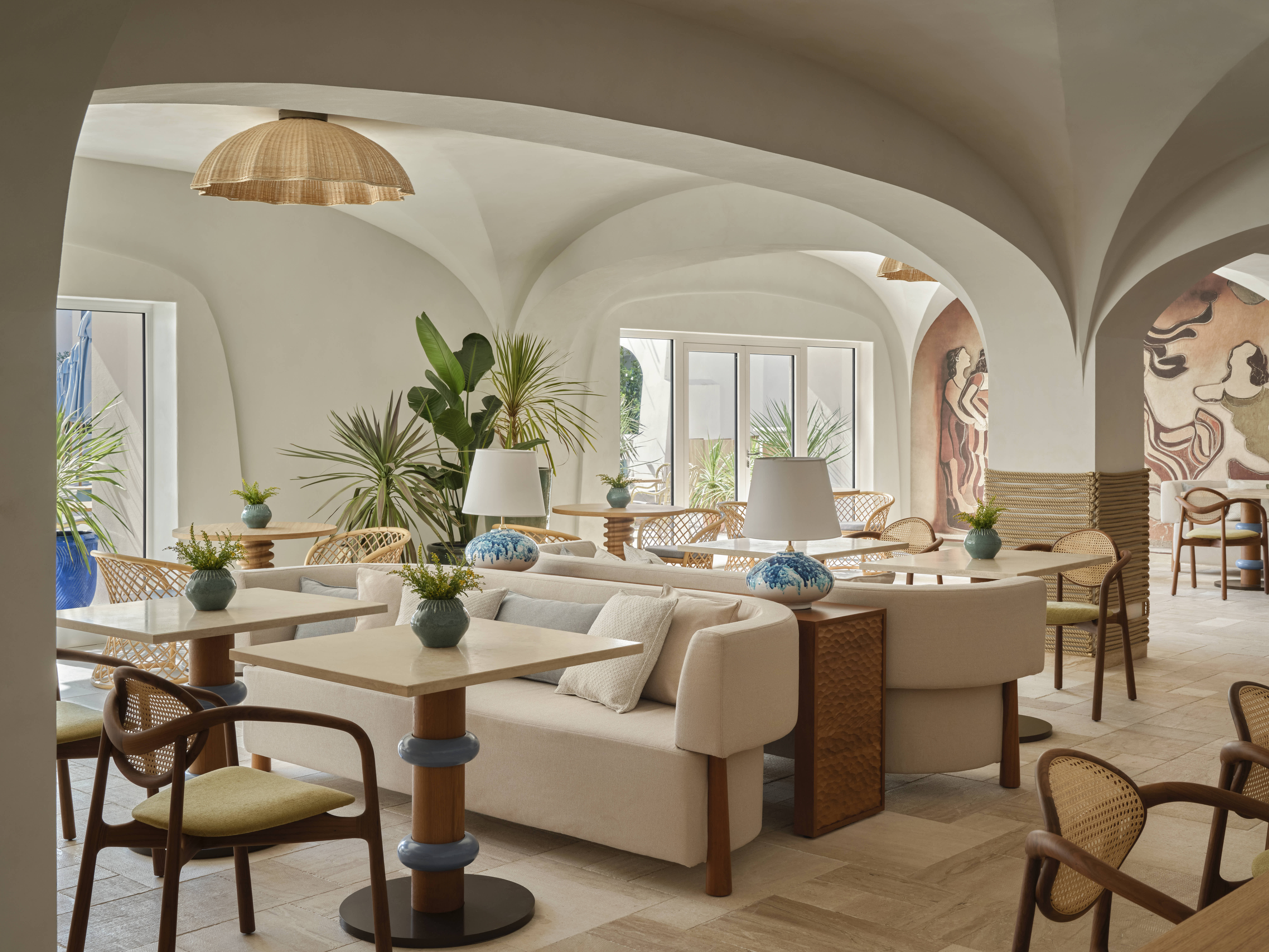 A seaside resort features spectacular, arched ceilings painted in a creamy tone, rattan and wooden furniture in playful silhouettes, turquoise accents, and a panoramic pool.