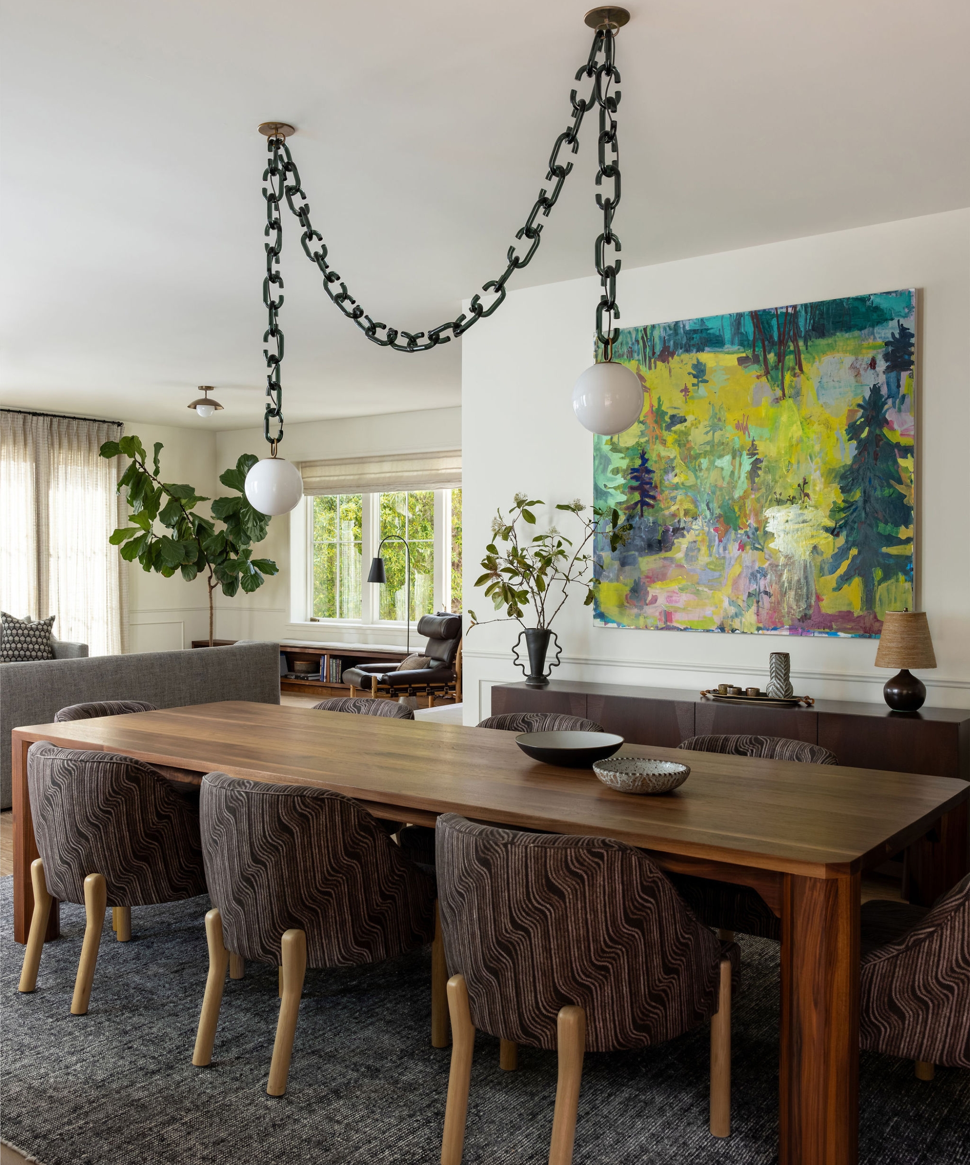 Dining room with a wood table, patterned upholstered chairs, a glass-chain pendant and a colorful landscape painting.