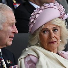 Queen Camilla wears a pink hat and pink suit jacket with a cream scarf and looks tearfully at husband King Charles