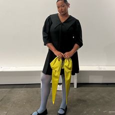 Imani wears black dress blue tights and black flats while holding a chartreuse bow bag