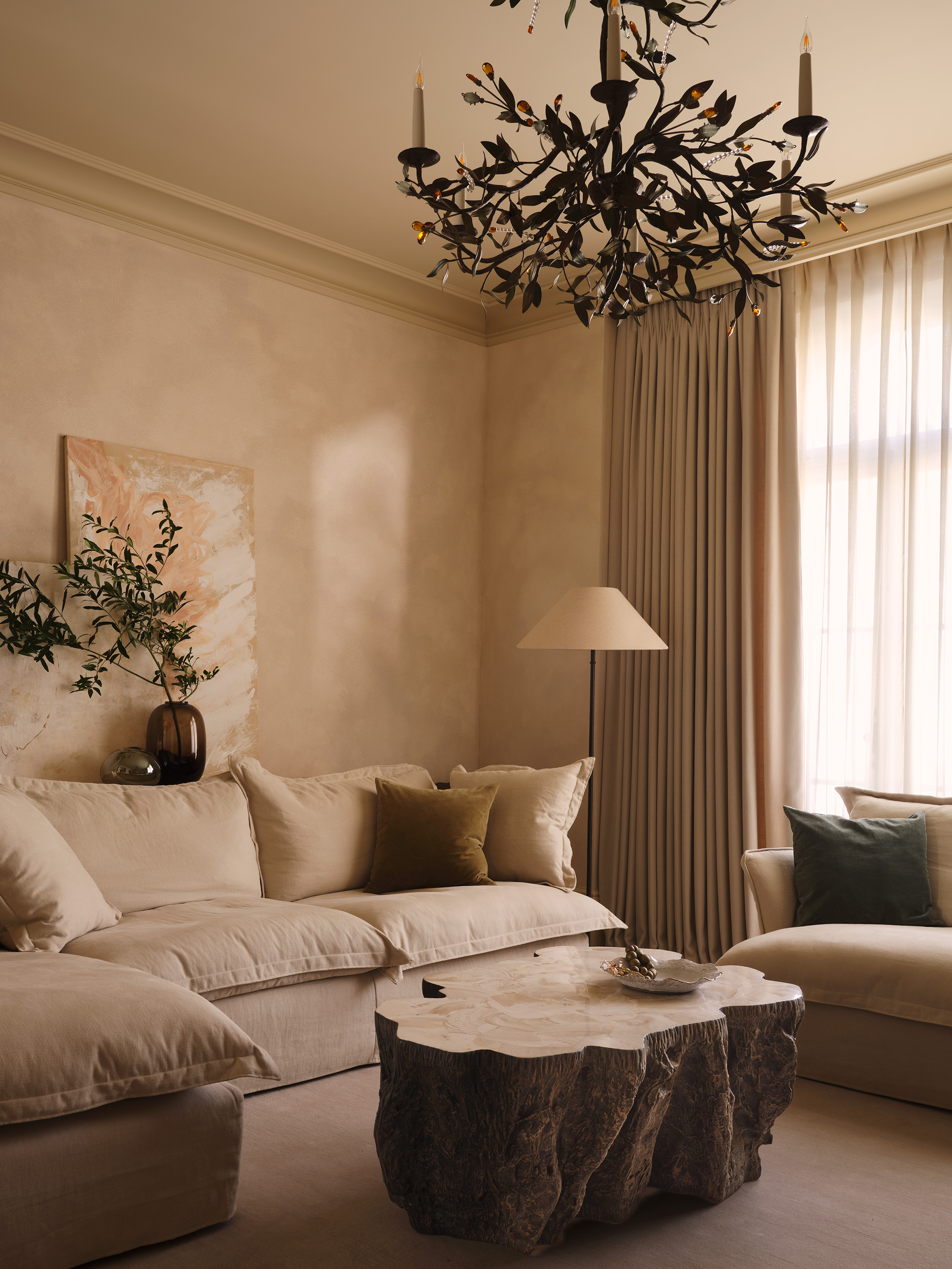 A living room with an abstract coffee table in the centre with a decorative dish on top underneath a leaf-like chandelier in a beige space