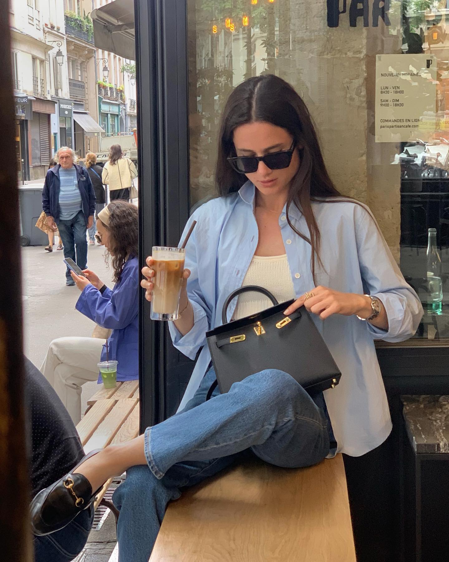 French fashion influencer sits in a café with an iced coffee wearing a blue button-down shirt, white top, Hermes bag, jeans, and Gucci loafers