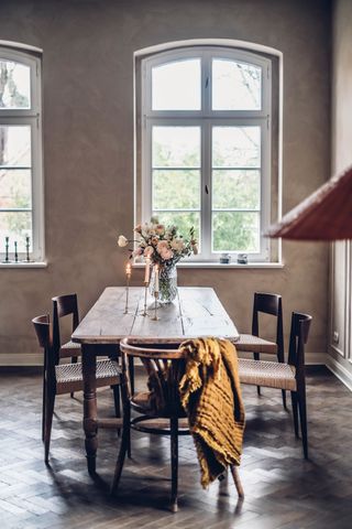 A long wooden farmhouse table featuring a a vase of flowers and three candlesticks by a large window