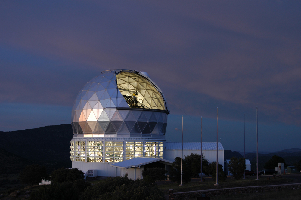 The mirror of the 9.2-meter Hobby-Eberly Telescope is visible through the open louvers in this twilight view. In daylight, the flagpoles on the right show the flags of the five HET partner institutions. 