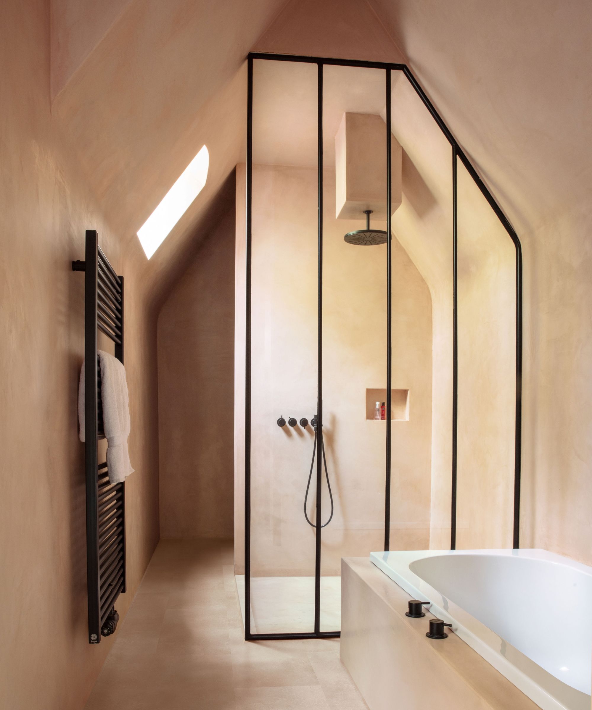 A sculptural attic bathroom finished entirely in warm blush-toned plaster, with a sloped ceiling and roof light. A black-framed glass shower enclosure houses a ceiling rain head and matte black controls, beside a built-in white bath with black taps and a ladder-style black heated towel rail.
