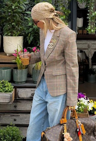 A woman wearing a plaid blazer with brooches, a white t-shirt, baggy jeans, a Fendi headscarf, and a Louis Vuitton bag with bag charms.