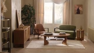A warm, contemporary living room with neutral walls and sheer curtains framing a large window. It features a green velvet loveseat, caramel leather chair, and oak furniture on a soft cream rug.