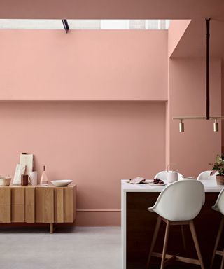 Pink painted kitchen, gray flooring, low, light wood sideboard, marble waterfall kitchen island, white and light wood bat chairs, skylight on ceiling
