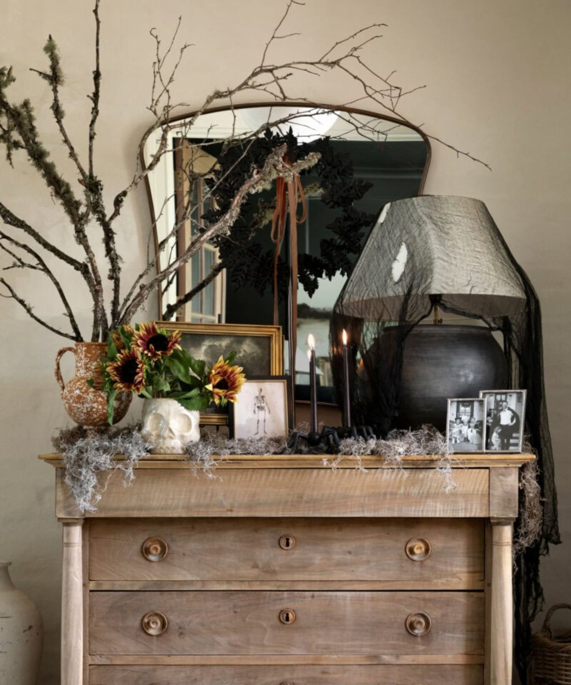 rustic chest of drawers styled for halloween with a mirror, rustic twigs, a lamp covered in black lace, cobwebs and a skull vase