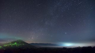 several meteors streak across the star-studded sky leaving long white trails across the night sky above a vegetated terrain.