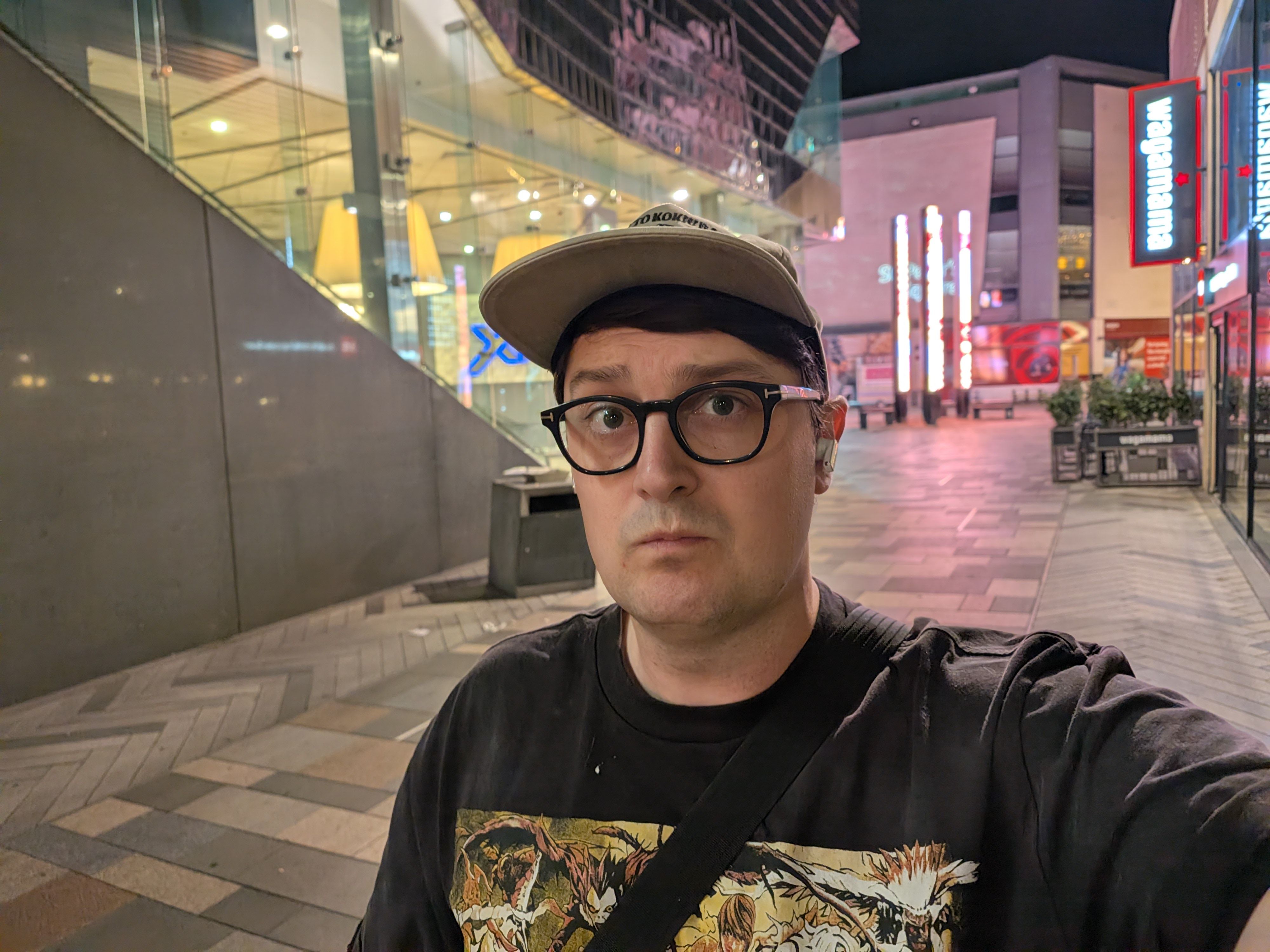 Man wearing glasses and a baseball cap taking a nighttime selfie on a pedestrian shopping street with illuminated signs behind him.