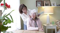 A woman brushes her mother's hair while she sits at a table in her bedroom.