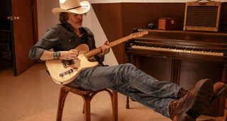 David Rawlings reclines on a chair in front of a piano and picks some notes on his Fender Telecaster. He wears a wide-brimmed hat.