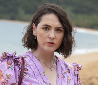 Amelia Templeton (Emma Sidi) stands on the beach with the ocean in the background. She is looking into the camera, looking faintly concerned.