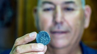 Jacob Sharvit (clean shaven, short white hair), director of the Marine Unit of the Israel Antiquities Authority, holds the up the Luna coin in his fingers. It’s an old coin with the profile of Luna, ancient goddess of the moon. Below her face there is the cancer zodiac sign.