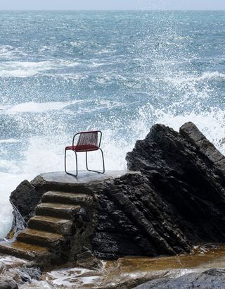 A photo of a red open-backed chair on a rock with waves from the sea rising up.
