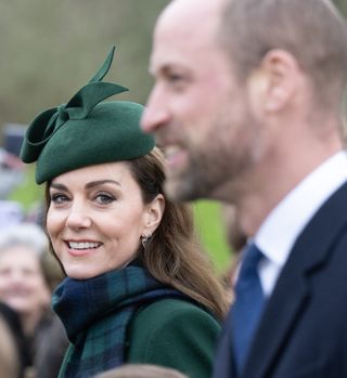 Kate Middleton wearing a green coat and plaid scarf with a green hat looking at Prince William, who is blurred out in the photo, and smiling