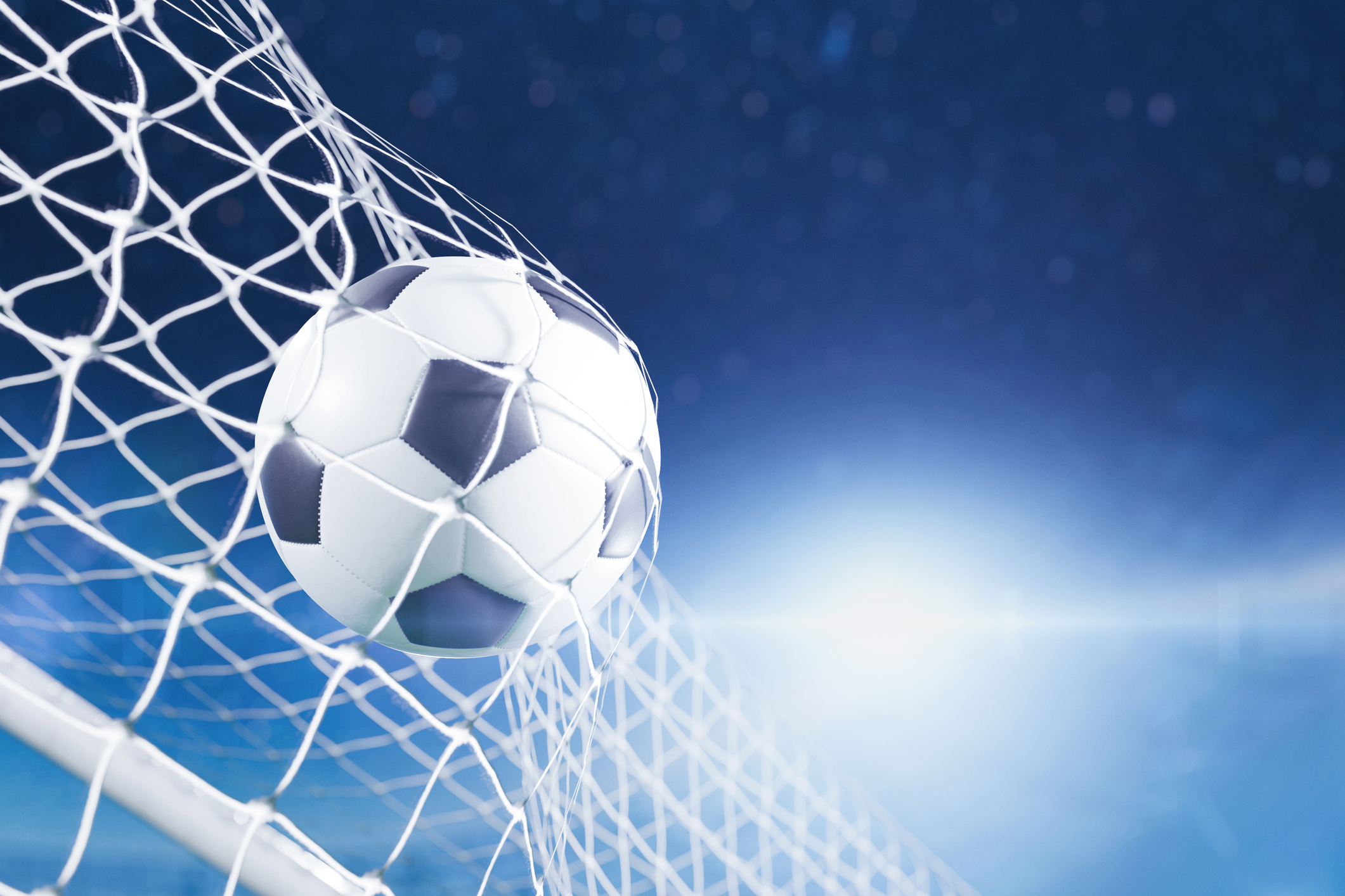 Close-up of a soccer ball hitting the net with a bright lens flare on a blue stadium background.