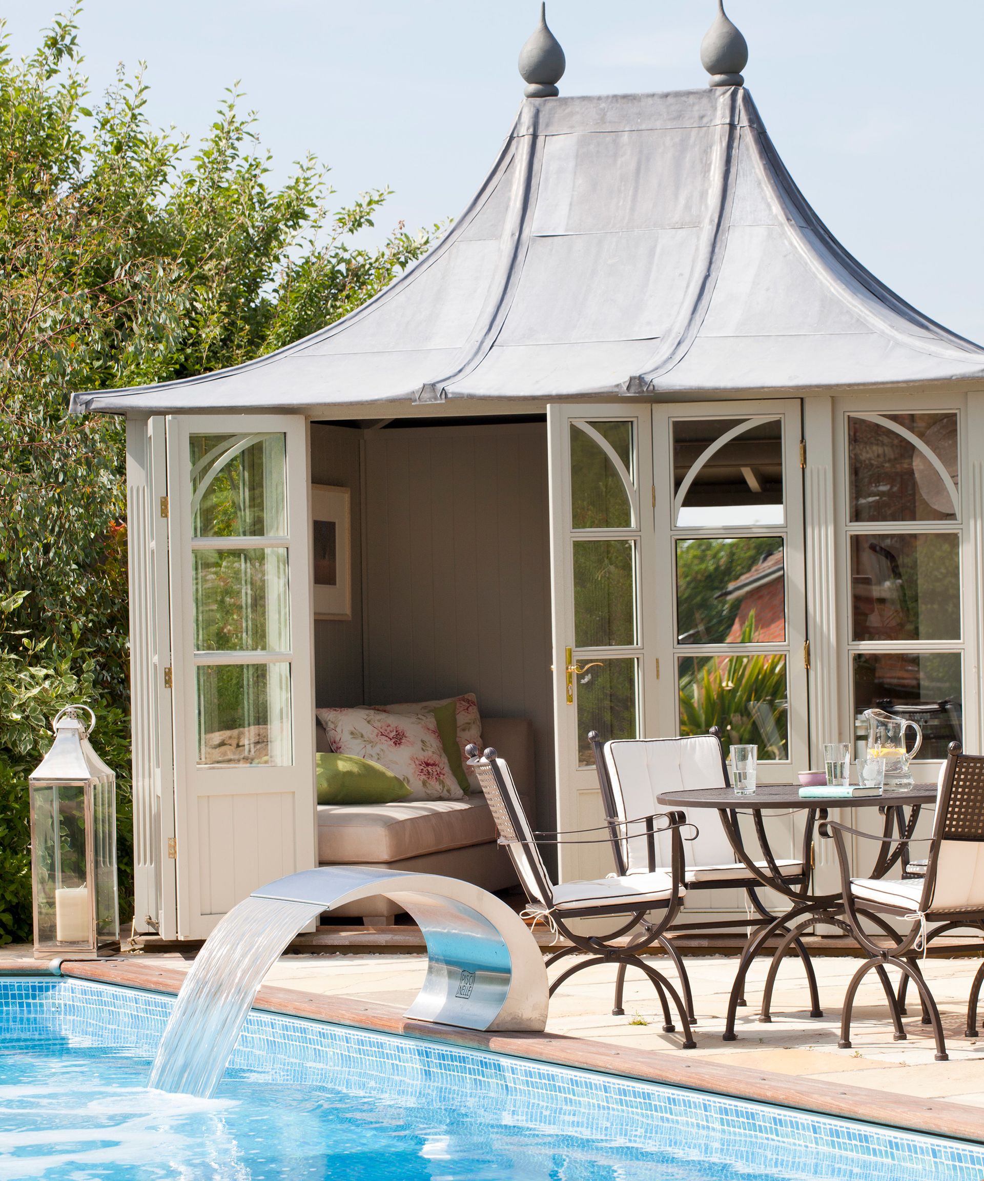 An example of pool party ideas showing a summer house, a dining table and a pool with a water feature
