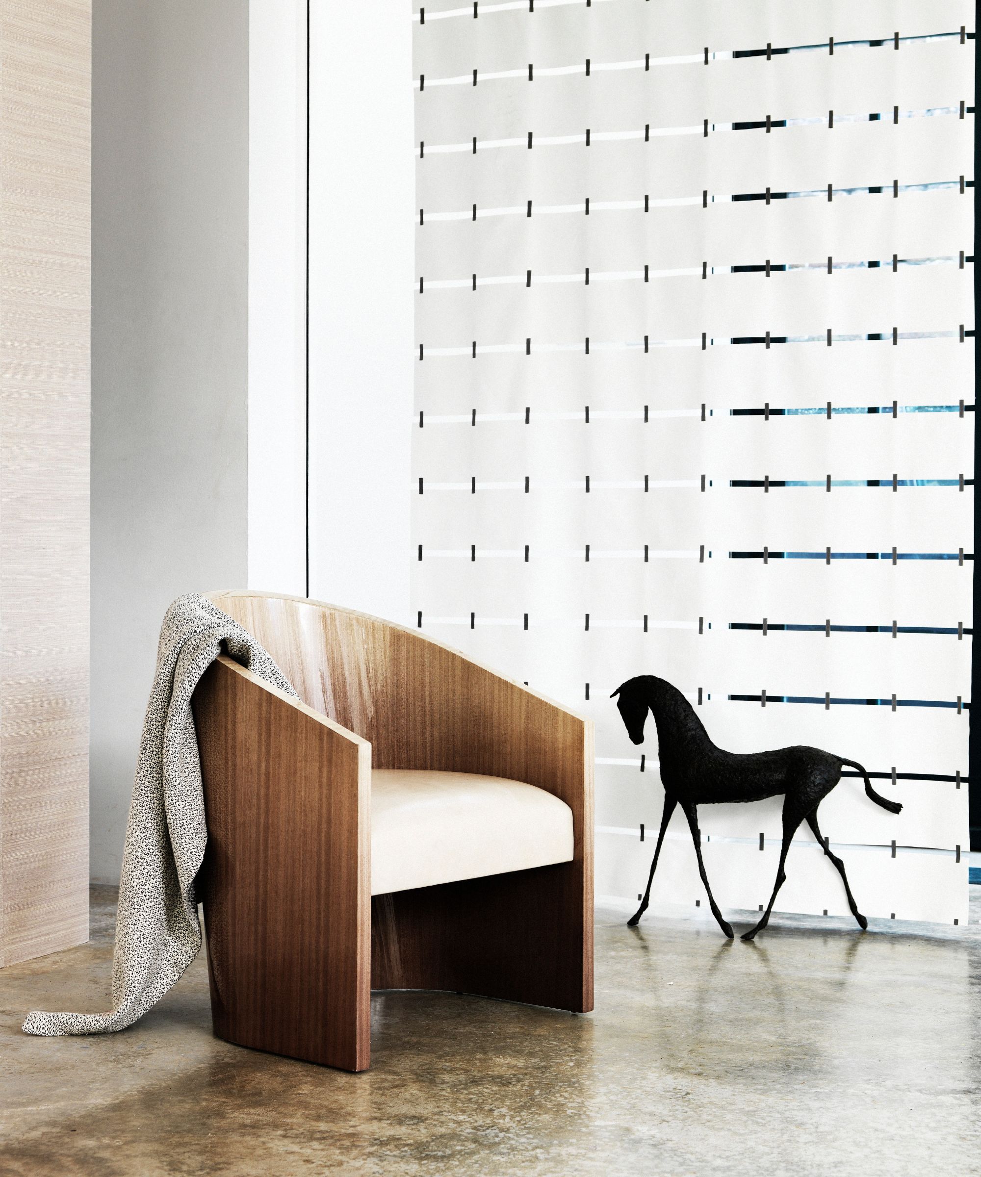 A minimalist wooden chair with grey blanket, black horse statue, marble floors and glass wall.