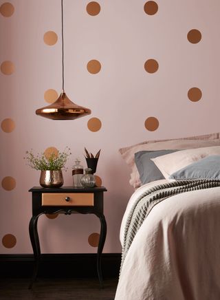 A pink bedroom with walls painted in Pashmina with copper spots by Crown, with a black bedside table and a pendant light