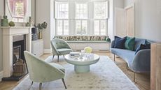 A bright living room with hardwood floors, white walls, and large windows with open white shutters. In the center of the room is a grey crushed velvet rug with a mint green and white circular coffee table and two matching mint green crushed velvet chairs. To the right is a blue velvet couch with blue and green cushions, and to the left is a white mantelpiece with a log burner, wicker baskets, and assorted vases.